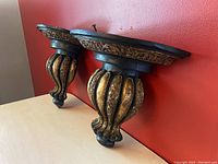 Side angled view of two ornate wall sconce shelves displaying curved bulbous base with gold and black finish.