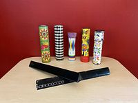 Six vintage kaleidoscopes arranged upright on a wooden surface against a red wall, showing colorful folk art, geometric, floral, and character designs on cylindrical tubes, plus a black flat rectangular box.