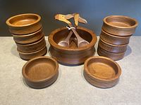 Photo showing the full teak salad bowl set with 10 small bowls stacked on either side of one large bowl with two bird-headed wooden serving tongs in the center.