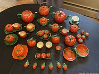 Full view of the 52-piece vintage tomato themed ceramic set arranged on a black surface, showing variety of teapots, pitchers, cups, salt and pepper shakers, and condiment holders in red with green leaves.