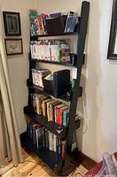 Full view of ladder-style bookcase with five painted dark wood shelves filled with books, DVDs, CDs, and other small items.