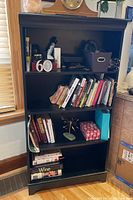 Full view of the black composite material bookcase showing four shelves filled with various items for context. Contents not included in sale.