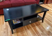 Black composite coffee table with rectangular top, curved legs, two open shelves, and one drawer with metal pull, shown at an angle.