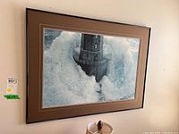Wide shot of the framed lighthouse print on a white wall showing the crashing waves and brick lighthouse, brown frame and matting.