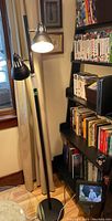 Full view of black metal floor lamp with two adjustable light sources beside bookshelf, showing lamp base and pole.
