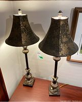 Pair of two table lamps on wooden surface, both lamps lit, showing bell-shaped floral fabric shades and ornate mixed material bases.