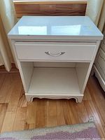 Front view of the white vintage nightstand showing drawer with silver handle and open shelf below.