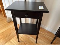 Side view of black wooden side table showing drawer and lower shelf, placed on light hardwood floor near white door and wall.