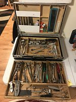 Open briefcase showing two wooden stacking trays filled with various vintage tools and small items organized in compartments.