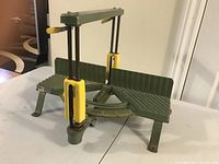 View of green cast iron mitre box with yellow clamps on a white table, showing vertical saw guides and angled base.