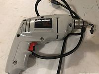 Side view of gray Duramark electric drill with power cord coiled beside it, showing the red trigger and metal chuck.