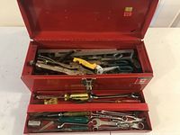 Open red metal tool box showing internal compartments with assorted hand tools inside including pliers, wrenches, screwdrivers, clamps, and scissors.