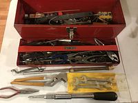 Top view of open red metal toolbox showing a variety of tools including vintage British wrenches, screwdrivers, and pliers in the central compartment with yellow package of wrenches and silver rod in front.