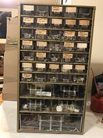 Full view of beige metal wall-mountable cabinet with 40 clear plastic drawers. Each drawer has a handwritten label describing contents including nuts, bolts, and faucet washers.