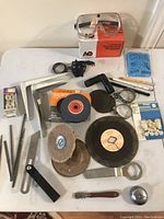 Full view of all miscellaneous tools arranged on a white surface including grinding wheel, protective goggles in box, wire brush wheels, wrench, electrical outlets, pencil, round discs and other hand tools.