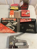 Wide view of the staple gun in original box and multiple boxes of staples mostly full, various brands and sizes visible.