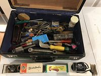 Open black suitcase with blue lining filled with assorted hand tools including wrenches, scrapers, and paintbrushes; three boxed items and Skil charger unit placed in front of the suitcase.