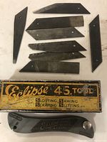 Top view showing 8 different metal blades arranged on a surface with the vintage Eclipse 4S tool handle near the bottom and the original metal box partially visible underneath.