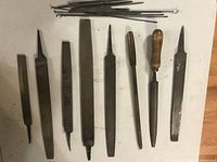 Top-down image showing 10 files and rasps of different shapes and sizes including some with wooden handles, laid out on white table surface with some long thin metal rods above them.