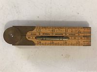 Photo of wooden folding measuring stick in the folded flat position showing the measurement scales, including inches and centimeters marked on wood with brass hinge and hardware details visible.