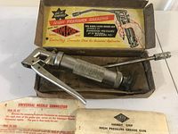 Metal grease gun laying inside original cardboard box with printed label. Instruction sheet partially visible alongside the gun.