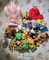 Image showing a collection of assorted baby toys including plush animals, character toys, and a baby car seat cover arranged on a floral surface.