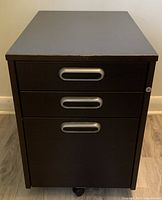 Front view showing all three drawers and silver handles, black finish visible. One caster wheel present at bottom right.