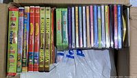 Photo showing a cardboard box containing multiple children's DVDs including Thomas & Friends Volumes 6 to 8, Hello Kitty DVDs, Pooh & Friends DVDs, Hollywood Classics DVDs, various children's music and cartoon CDs, and three Visual VHS tapes.