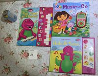 Top down view of three children's interactive music books featuring Barney and Dora The Explorer characters, each with built-in piano keyboards or musical keypads, placed on floral fabric background with a soda can for size reference.
