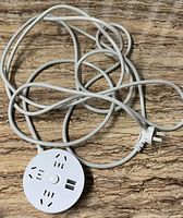 Photo of the round white power outlet extension showing three universal-type sockets, power switch, two USB ports, attached white cable and plug.