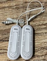 Photo showing two white oval electric shoe dryers with ventilation holes, connected by cords with a plug, placed on a textured surface.