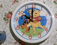 Front view of round clock with white frame and Winnie the Pooh and Tigger dancing on blue sky background, red numbers around