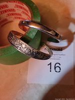 Two 925 silver bangle bracelets shown with a roll of green tape as background. One bracelet is plain polished silver, the other is engraved with floral and pattern designs.
