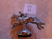 Photo of a silver-tone antique metal hood ornament shaped as a galloping horse with a jockey on its back, showing signs of pitting due to age.