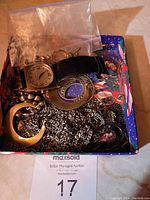 Top view of a box filled with assorted broken jewelry pieces and a wristwatch, showing different chains, pendants, and earrings in a colorful box.