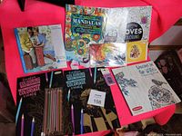 Photo showing coloring books, colored pencils, Prismacolor pencils, and pencil sharpener on a pink fabric background.