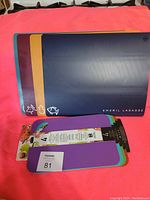 Photo showing two sets of cutting mats on a flat surface. The large set has four mats in blue, yellow, purple, and dark blue, with the dark blue mat branded Emeril Lagasse and showing small white vegetable and fish icons. The small set comprises four purple and green mats still in packaging, labeled Easy Ratchet Cutting Board.