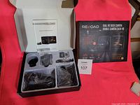 Opened Reload dual HD dash camera box showing main camera, accessories, cables and memory cards, with retail box beside it on pink background.