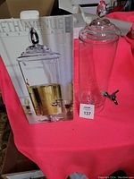 Photo of the clear glass drink dispenser next to its original box on a pink cloth background.