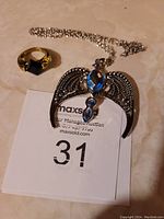 Silver-tone necklace with ornate wings and blue stones alongside gold-tone ring with amber-toned crystal on a neutral background with auction tag.