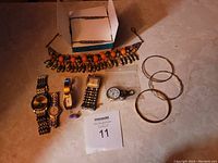 Necklace with orange beads, multiple wristwatches, and three metal bangle bracelets shown on floor with lot tag 11