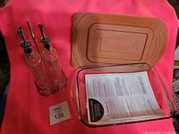 Photo of Rachael Ray glass baking dish with brown silicone lid, and two glass oil and vinegar bottles with metal pour spouts sitting in a metal rack, placed on a red surface.
