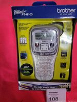 Photo of unopened Brother P-Touch PT-H100 label maker box. The label maker is handheld with QWERTY keys and a display. Packaging is yellow and purple with branding and usage info.