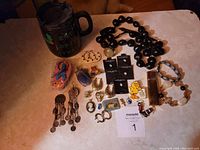 Photo showing a collection of costume jewelry consisting of a chunky black bead necklace, assorted earrings, bracelets, rings, brooches, and a black travel mug with designs.