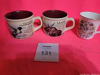 Three ceramic soup mugs on a pink background with auction tag 131. Two mugs feature wildlife illustrations (Ruby-throated Hummingbird and Moose), and one smaller mug depicts a royal couple.