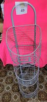 Photo of the three-tier metal wire storage basket unit with oval baskets and a handle, positioned against a pink background.
