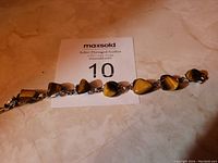 Photo showing a linked bracelet with multiple tiger's eye stones shaped as small hearts and ovals connected by silver-toned metal links, placed on a beige surface