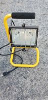 Front view of a portable worklight with yellow metal frame, protective grill, foam handle, and power cord on asphalt ground.