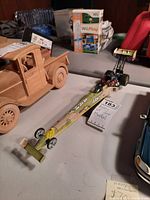 Photo showing the full length of the dragster model car on a table with a small tag stating 'Army of one Model'. Adjacent wooden car model is partially visible.