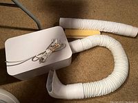 White rectangular BedJet unit with attached power cord and two flexible white air hoses on carpeted floor.